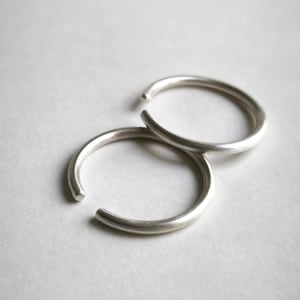 Könnte beinhalten: Zwei silberne Ringe, jeder mit einer einfachen, abgerundeten Form, liegen übereinander. Die Ringe sind aus glattem, poliertem Metall.