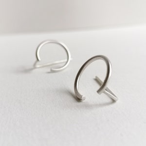 Peut inclure: Deux boucles d'oreilles minimalistes en argent avec un design circulaire. Les boucles d'oreilles sont faites d'une seule pièce de métal qui est pliée en un cercle avec une petite barre qui s'étend du cercle.
