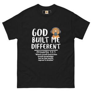 Könnte beinhalten: Schwarzes T-Shirt mit dem weißen Text "GOD BUILT ME DIFFERENT" und einer Cartoonfigur mit Kopfhörern. Das T-Shirt enthält auch einen Bibelvers aus Sprüche 12:1.