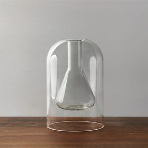 May include: A clear glass vase with a distinctive design. It has a bell-shaped outer layer and an inner flask-shaped vessel. The transparent glass allows a view of the interior. The vase is placed on a wooden surface.
