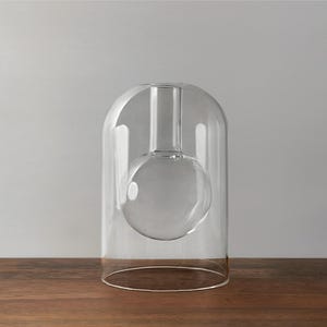May include: A clear glass vase with a spherical base and a cylindrical neck, enclosed within a larger, arched glass structure. The vase rests on a wooden surface against a neutral grey background. The design is minimalist and modern.