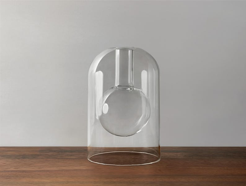 May include: A clear glass vase with a spherical base and a cylindrical neck, enclosed within a larger, arched glass structure. The vase rests on a wooden surface against a neutral grey background. The design is minimalist and modern.