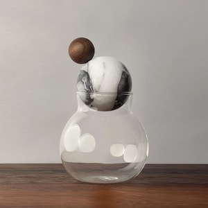 May include: A clear glass vase with a marble and wood ball-shaped lid. The marble ball has gray and white veining, and the wood ball is a dark brown color. The vase sits on a dark brown wooden surface.