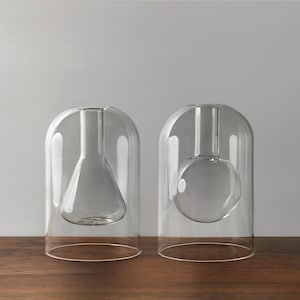 May include: Two clear glass vases with a modern design. One vase has a conical interior, and the other has a spherical interior. Both are encased in a rounded, bell-shaped glass exterior. The vases sit on a dark wooden surface.