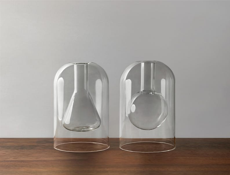 May include: Two clear glass vases with a modern design. One vase has a conical interior, and the other has a spherical interior. Both are encased in a rounded, bell-shaped glass exterior. The vases sit on a dark wooden surface.