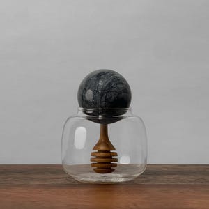 May include: A clear glass honey jar with a black marble lid and a wooden honey dipper. The dipper has a honeycomb design.