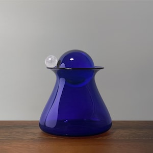May include: A deep blue glass jar with a spherical lid and a small clear crystal ball on top. The jar has a rounded base and a flared opening. The jar is on a wooden surface.