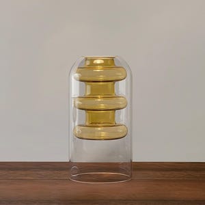 May include: A clear glass vase with three stacked, amber-colored glass rings. The vase is on a wooden surface.