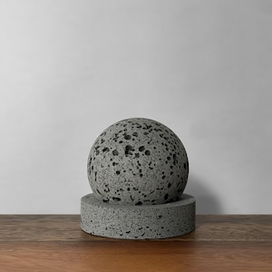 Può includere: Una sfera di pietra grigia porosa è posta sopra una base cilindrica abbinata. Gli oggetti hanno una superficie ruvida e strutturata e sono esposti su una superficie di legno su sfondo bianco. La sfera ha un diametro di circa 10 cm.