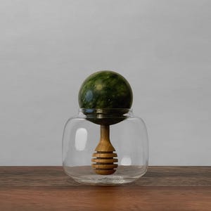Op de afbeelding: Een heldere glazen pot met een houten honingdipper en een groene marmeren bol erop. De honingdipper is gemaakt van lichtbruin hout en heeft een spiraalvormig ontwerp. De pot staat op een houten oppervlak.