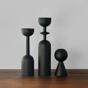 May include: Three black wooden candle holders of varying heights and shapes. The tallest has a cup-like top, the middle one has a rounded top, and the shortest has a spherical top. All are displayed on a wooden surface against a white background.