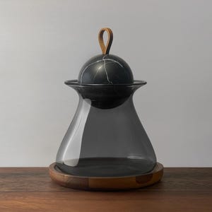 May include: A decorative glass jar with a dark gray tint, a black marble lid, and a brown leather loop handle. The jar sits on a round wooden base, perfect for storing items.