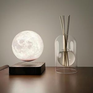 May include: A spherical moon lamp, appearing to float above a dark wooden base. Next to it, a clear glass diffuser with reeds, creating a modern and minimalist aesthetic. The lamp and diffuser are displayed on a wooden surface.