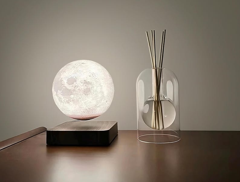 May include: A spherical moon lamp, appearing to float above a dark wooden base. Next to it, a clear glass diffuser with reeds, creating a modern and minimalist aesthetic. The lamp and diffuser are displayed on a wooden surface.