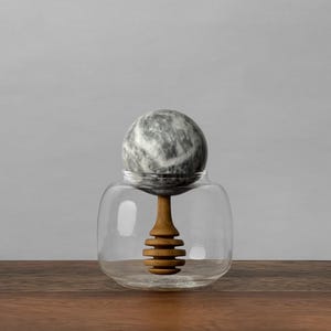 May include: A clear glass honey jar with a wooden honey dipper and a grey and white marble lid. The jar sits on a wooden surface against a grey background. The honey dipper is made of light brown wood.