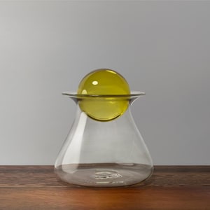 Puede incluir: Un jarrón de vidrio transparente con base redondeada y borde acampanado, coronado por una esfera de vidrio amarillo-verdoso. El jarrón está sobre una superficie de madera contra un fondo gris neutro. El diseño es moderno y minimalista.