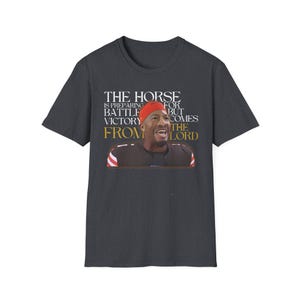 Pode incluir: Uma camiseta cinza escura com um gráfico de um jogador de futebol americano usando um capacete vermelho e uma camisa dourada. O texto na camiseta diz "The Horse is preparing for battle but victory comes from the Lord."