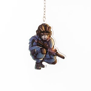 May include: A blue and black cartoon character wearing a helmet and holding a gun. The character is crouching and looking to the right. The character is printed on a clear acrylic keychain.