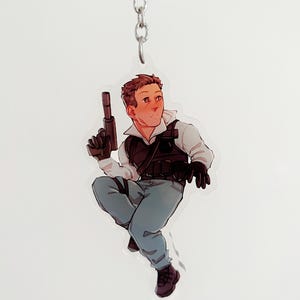 May include: A clear acrylic keychain featuring a cartoon illustration of a person in tactical gear, holding a gun. The person is wearing a black vest, gloves, and boots, with blue pants and a white shirt. The keychain hangs from a silver chain.