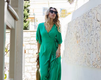 May include: A woman wearing a vibrant green wrap dress and sunglasses walks down a white staircase. She carries a woven handbag and wears beige heels. The dress has short sleeves and a flowing skirt, perfect for a summer day.