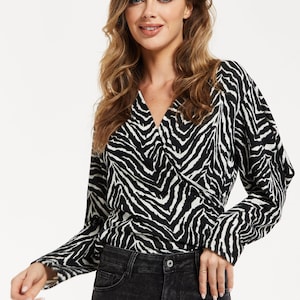 Pode incluir: Um top cruzado com estampa de zebra em preto e branco com mangas compridas e decote em V. O top é combinado com jeans pretos. A modelo tem cabelo loiro longo e ondulado.