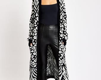 May include: A long, open-front cardigan in black and white animal print. The pattern combines zebra stripes and leopard spots. The cardigan is worn over a black top and black pants. The model is wearing black boots.