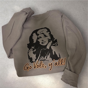May include: A taupe long-sleeve crewneck sweatshirt featuring a black graphic illustration of a person with voluminous hair. Below the graphic, the text "Go Vols, y'all!" is printed in orange lettering with a black outline on the front of the garment.