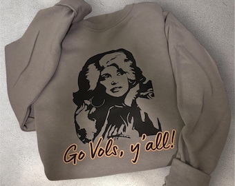 May include: A taupe long-sleeve crewneck sweatshirt featuring a black graphic illustration of a person with voluminous hair. Below the graphic, the text "Go Vols, y'all!" is printed in orange lettering with a black outline on the front of the garment.