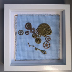 Puede incluir: Una obra de arte enmarcada que presenta un fondo de tela azul con una colección de engranajes de bronce y una esfera de reloj, junto con una llave.