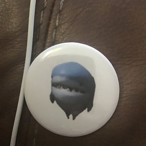 May include: A white button with a blue and grey dripping shark graphic.