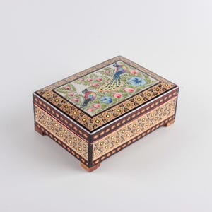 May include: A rectangular wooden box with four small feet, featuring a hand-painted lid with two colorful peacocks surrounded by pink and blue floral motifs. The sides of the box are decorated with intricate black, gold, and white geometric patterns.