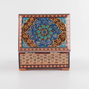 May include: A rectangular wooden box with a hinged lid featuring a vibrant blue, gold, and black floral mandala pattern. The base of the box is decorated with a repeating geometric pattern in shades of orange, black, and cream with small red accents.