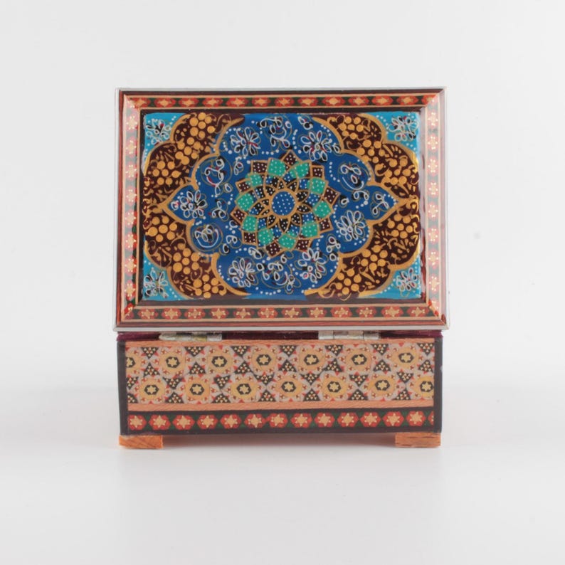 May include: A rectangular wooden box with a hinged lid featuring a vibrant blue, gold, and black floral mandala pattern. The base of the box is decorated with a repeating geometric pattern in shades of orange, black, and cream with small red accents.