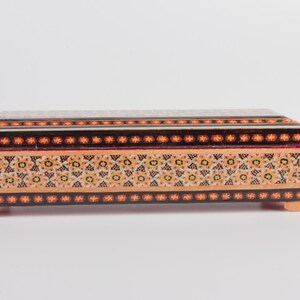 May include: A rectangular wooden box with intricate inlaid designs in shades of orange, yellow, black, and red. The box has a decorative border and rests on four small feet.