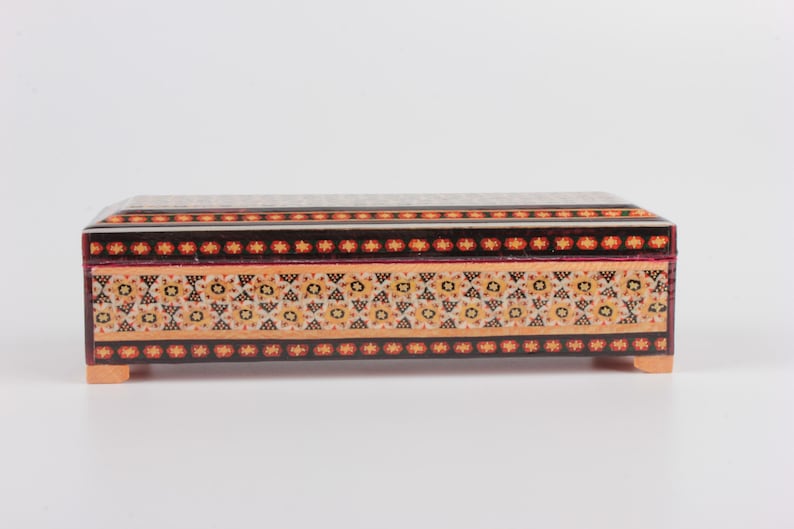 May include: A rectangular wooden box with intricate inlaid designs in shades of orange, yellow, black, and red. The box has a decorative border and rests on four small feet.