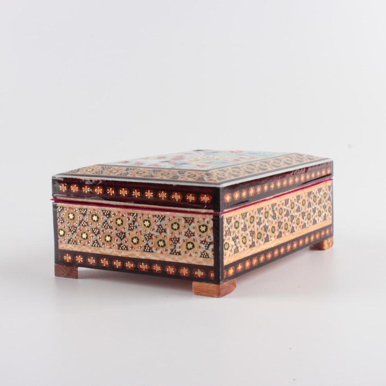May include: A rectangular wooden box featuring intricate geometric marquetry patterns in shades of black, cream, and gold. The box has a hinged lid, a smooth polished finish, and stands on four small, square wooden feet at each corner.