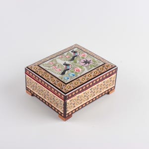 May include: A rectangular wooden box featuring intricate marquetry patterns on the sides and a lid decorated with a painted scene of two birds, pink flowers, and green foliage. The box is supported by four small, square wooden feet at the base.