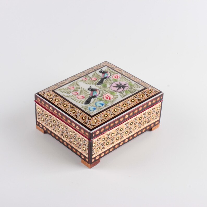 May include: A rectangular wooden box featuring intricate marquetry patterns on the sides and a lid decorated with a painted scene of two birds, pink flowers, and green foliage. The box is supported by four small, square wooden feet at the base.