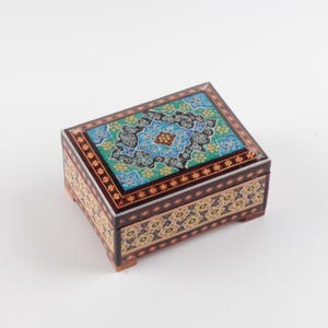May include: A rectangular wooden box featuring intricate floral and geometric patterns in shades of turquoise, blue, black, and gold. The lid displays a central medallion design, while the sides are decorated with repeating motifs and small feet.