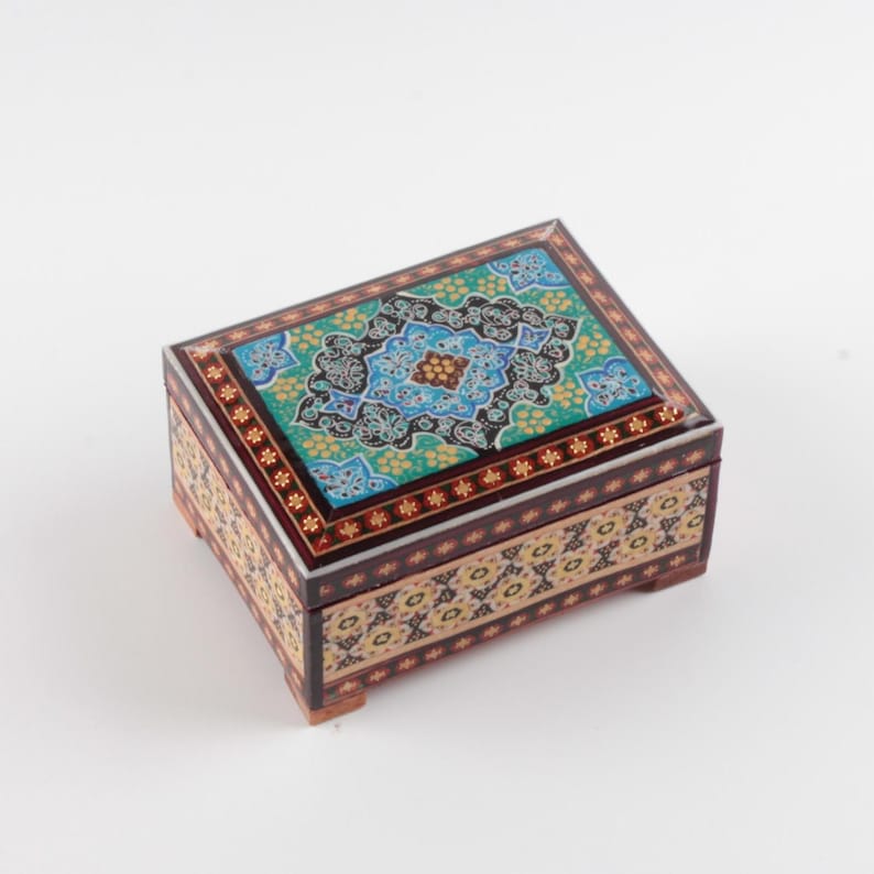 May include: A rectangular wooden box featuring intricate floral and geometric patterns in shades of turquoise, blue, black, and gold. The lid displays a central medallion design, while the sides are decorated with repeating motifs and small feet.
