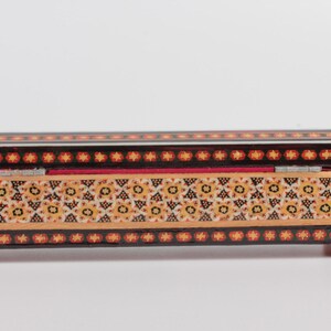 May include: A rectangular wooden box with a hinged lid. The box is decorated with intricate geometric patterns in shades of black, red, yellow, and white. The box has small feet on each corner.