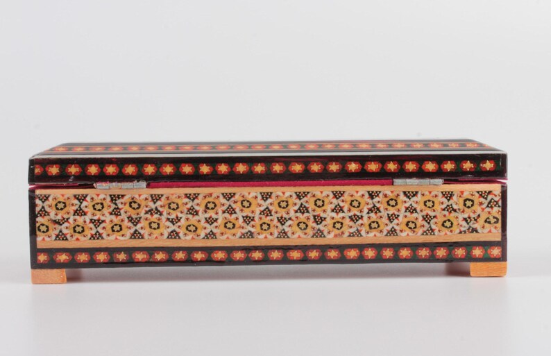 May include: A rectangular wooden box with a hinged lid. The box is decorated with intricate geometric patterns in shades of black, red, yellow, and white. The box has small feet on each corner.