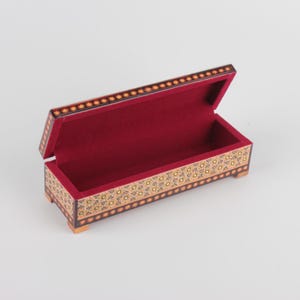 May include: A rectangular wooden box with a red velvet interior. The box is decorated with intricate geometric patterns in gold, black, and brown.