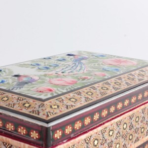 May include: A rectangular box featuring a lid with a painted scene of birds and pink flowers on a white background. The sides of the box are decorated with intricate geometric patterns in shades of brown, gold, red, and green, creating a detailed border.