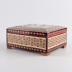 May include: A rectangular wooden box featuring intricate marquetry patterns in shades of brown, cream, and gold. The lid and sides are decorated with geometric star motifs and floral accents. The box is supported by four small, square wooden feet.