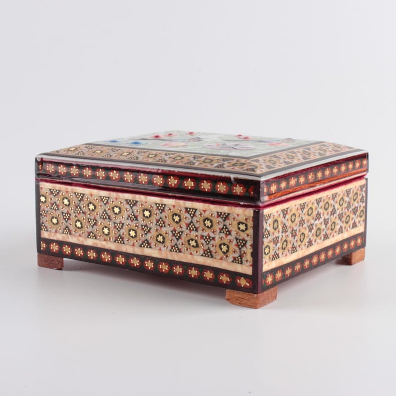 May include: A rectangular wooden box featuring intricate marquetry patterns in shades of brown, cream, and gold. The lid and sides are decorated with geometric star motifs and floral accents. The box is supported by four small, square wooden feet.