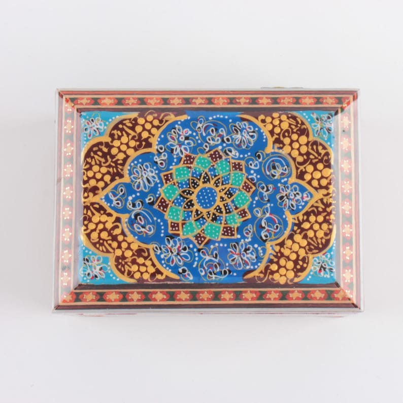 May include: A rectangular decorative box featuring a vibrant blue central medallion with a geometric floral pattern in turquoise, white, and gold. The surrounding border displays a brown and gold dotted motif, framed by a thin red and gold edge.