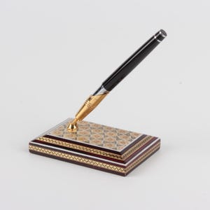 May include: A black pen with a gold-colored nib and accents rests in a decorative pen holder. The pen holder is rectangular with a patterned top and a dark wood base, featuring gold and cream-colored accents.