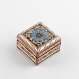 Könnte beinhalten: Eine quadratische Holzschmuckschatulle mit aufwendigen Intarsienmustern. Der Deckel zeigt ein zentrales blau-goldenes Blumenmuster, umgeben von schwarzen und goldenen Akzenten. Die Schatulle hat einen roten Innenraum und ist mit goldenen und cremefarbenen Details verziert.