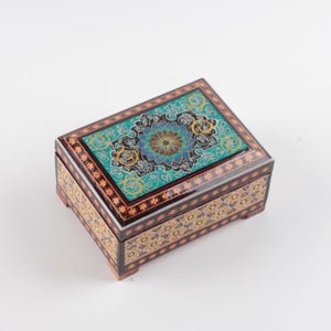 May include: A rectangular wooden box with a hinged lid featuring a central teal floral medallion surrounded by intricate black and gold patterns. The sides of the box are decorated with repeating gold floral motifs on a dark brown background.
